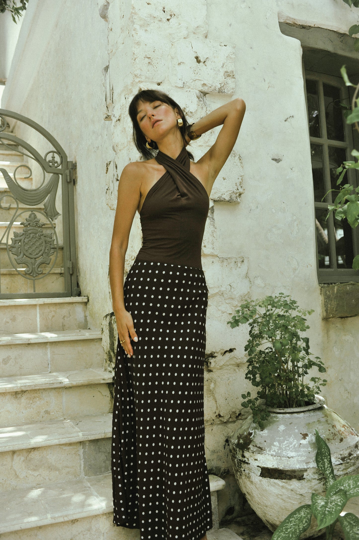 Woman in a brown top and black polka dot skirt standing against a white wall with plants.