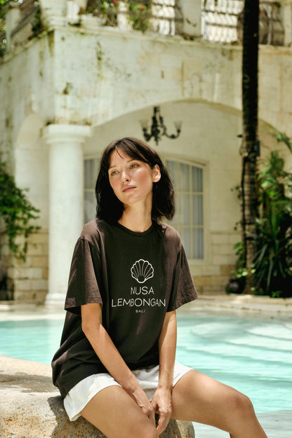Woman wearing a black t-shirt with 'Nusa Lembongan' text, sitting by a pool with a building in the background.