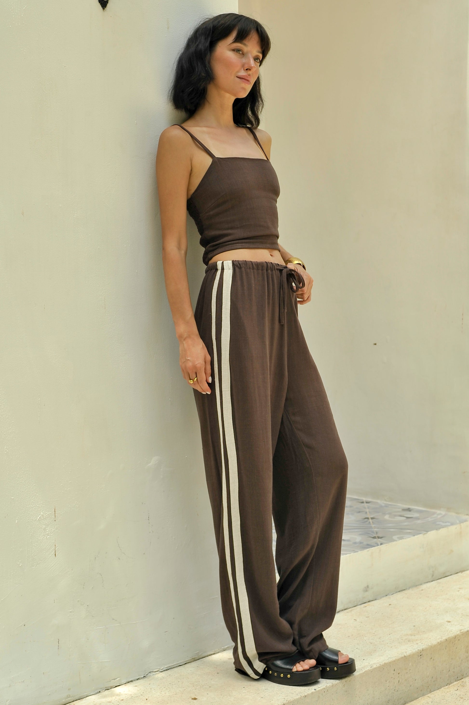 Woman wearing a brown top and wide-leg pants with white stripes against a light-colored wall.